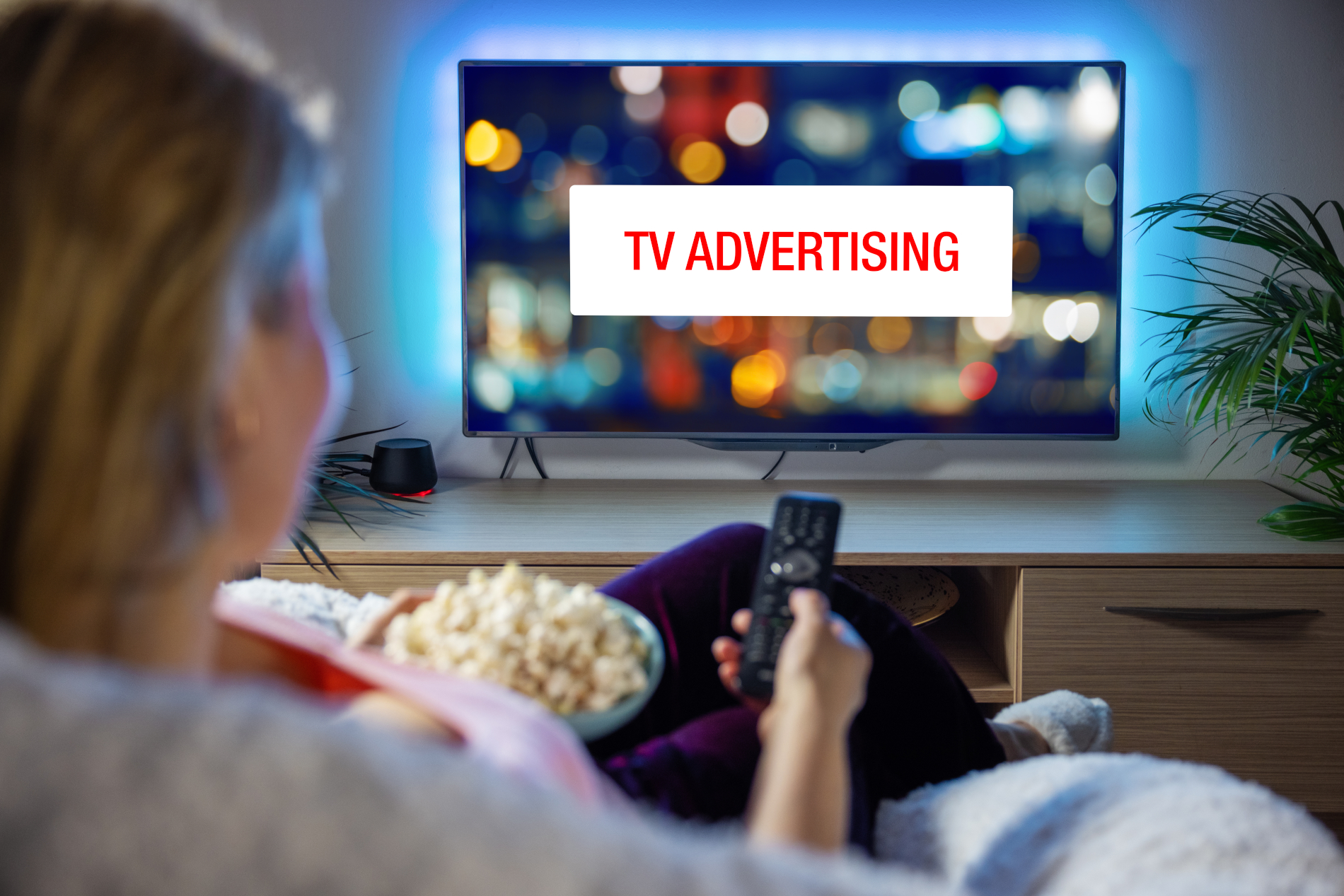 A woman sits at home with popcorn on her couch, pointing her remote towards a TV that says "TV Advertising" on it.