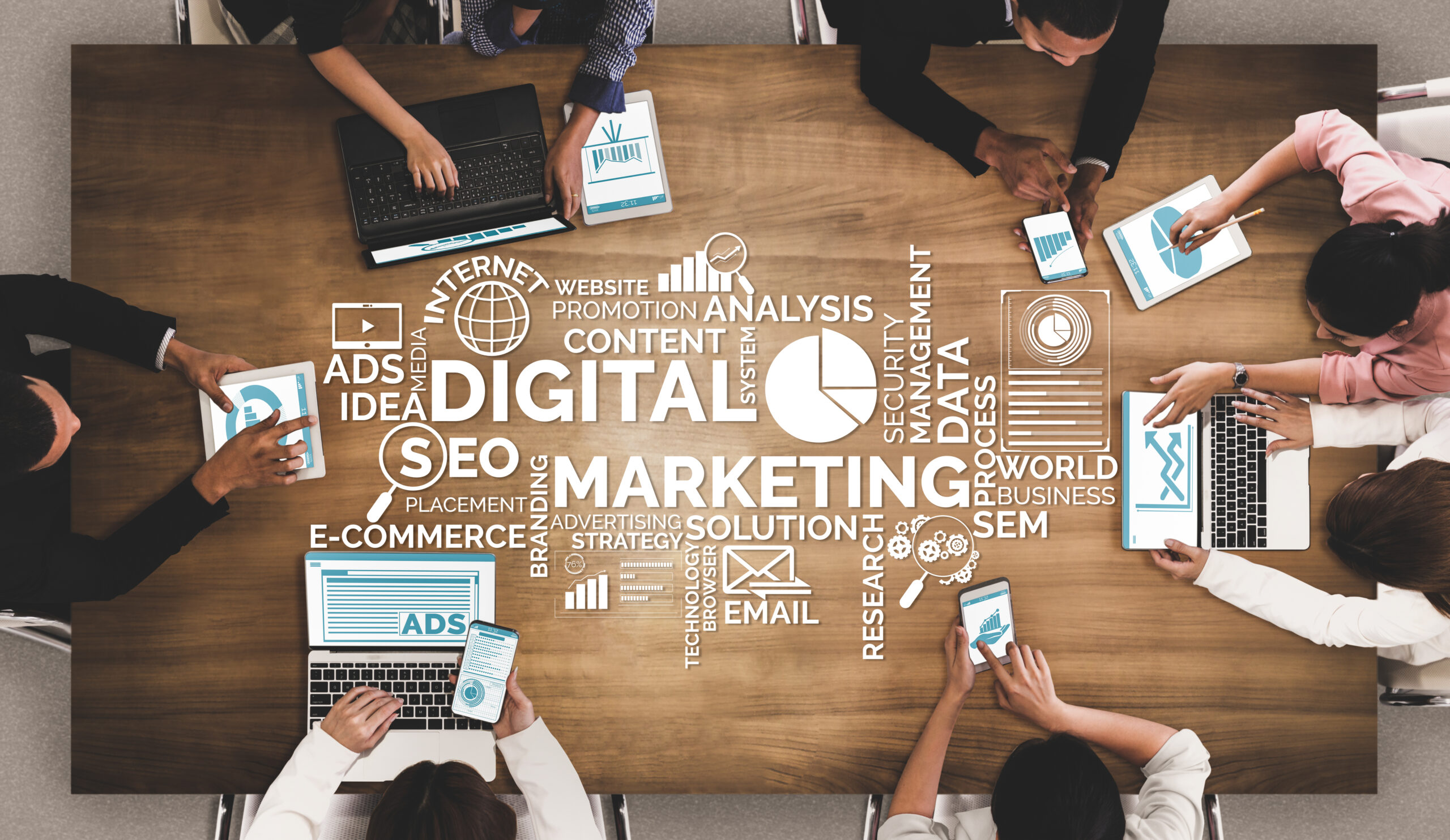 Seven digital marketers sit around a table and compare notes and charts. A graphical image including digital marketing buzzwords is layered over the image of the table