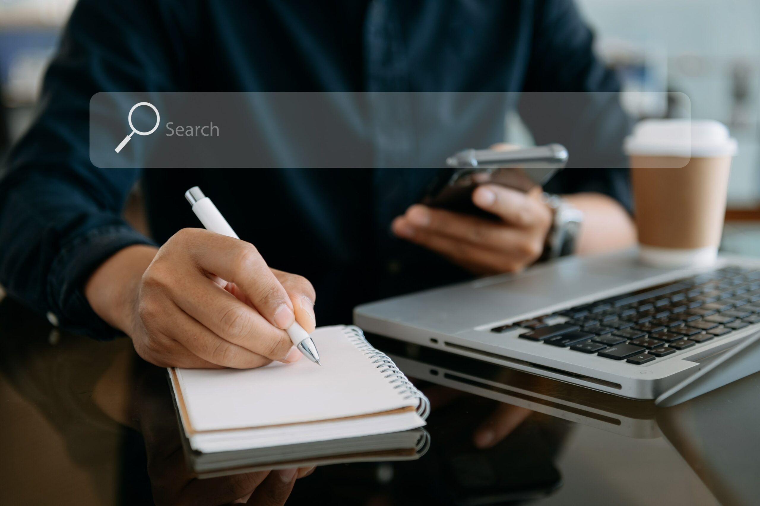 A user doing keyword research. They are seated at a desk writing information on a notepad while looking up information on a computer and smartphone.