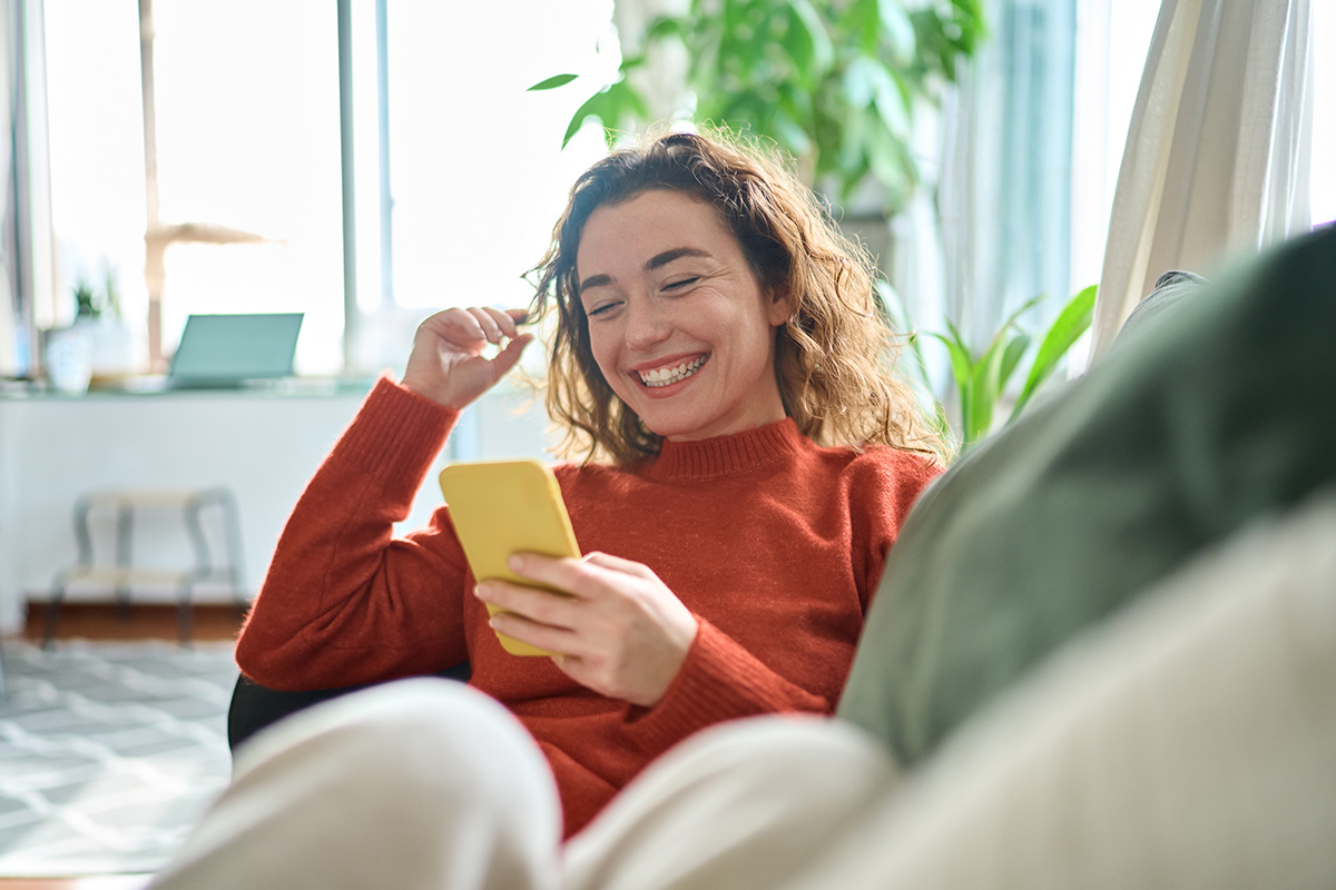 A woman smiles at her phone, sitting on a couch in a well lit room